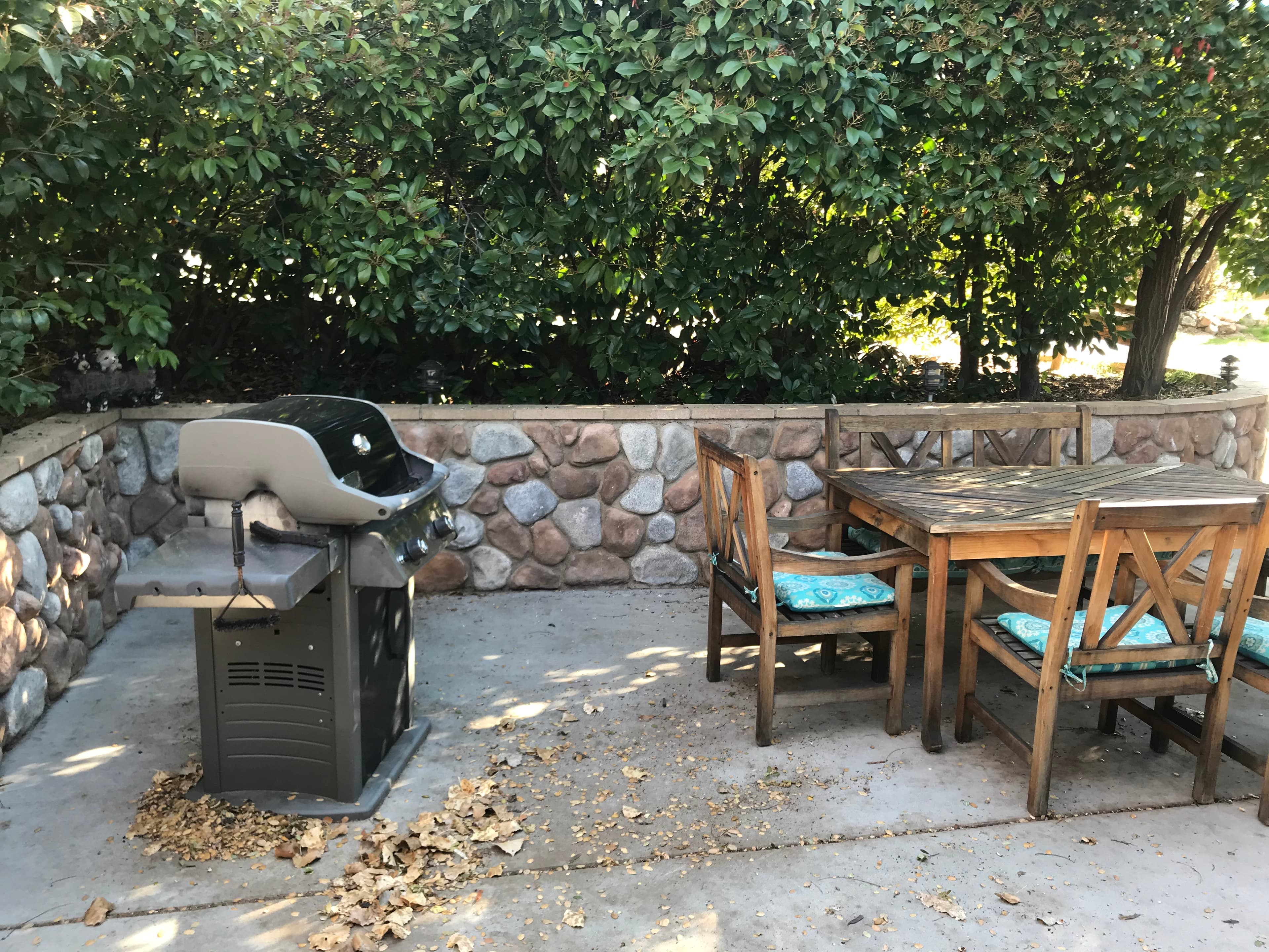 A gas grill next to a wooden dining table and chairs in a stone-walled outdoor patio.