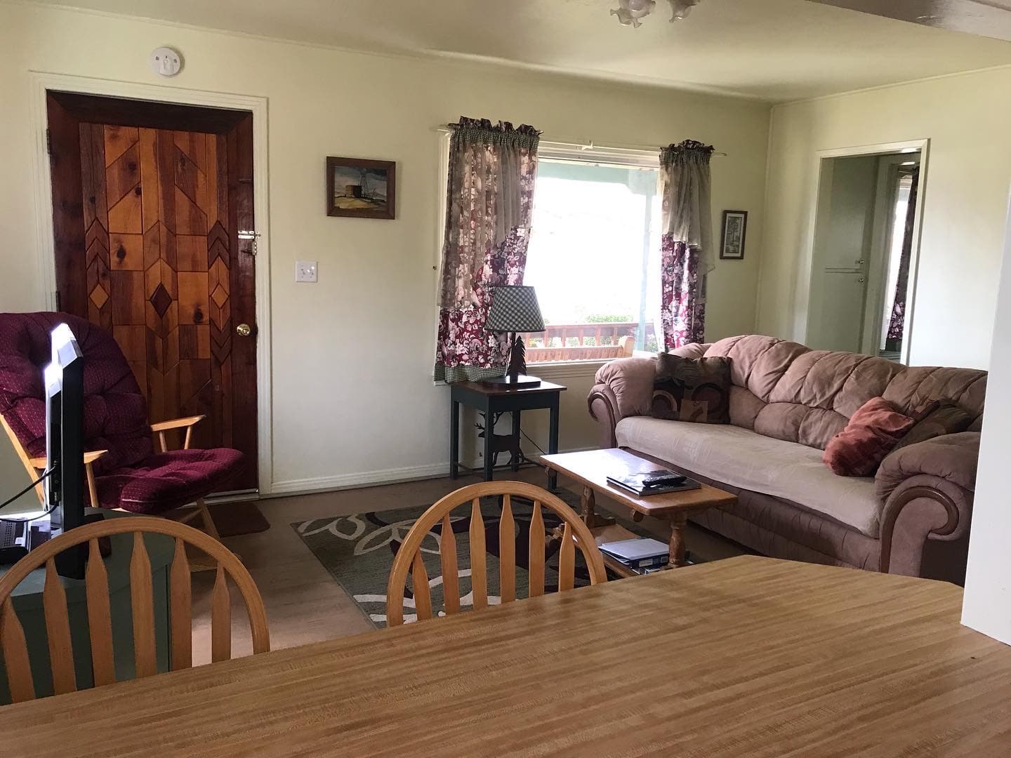 A cozy living room featuring a brown couch, a wooden table with chairs, a lamp, and a decorative front door.
