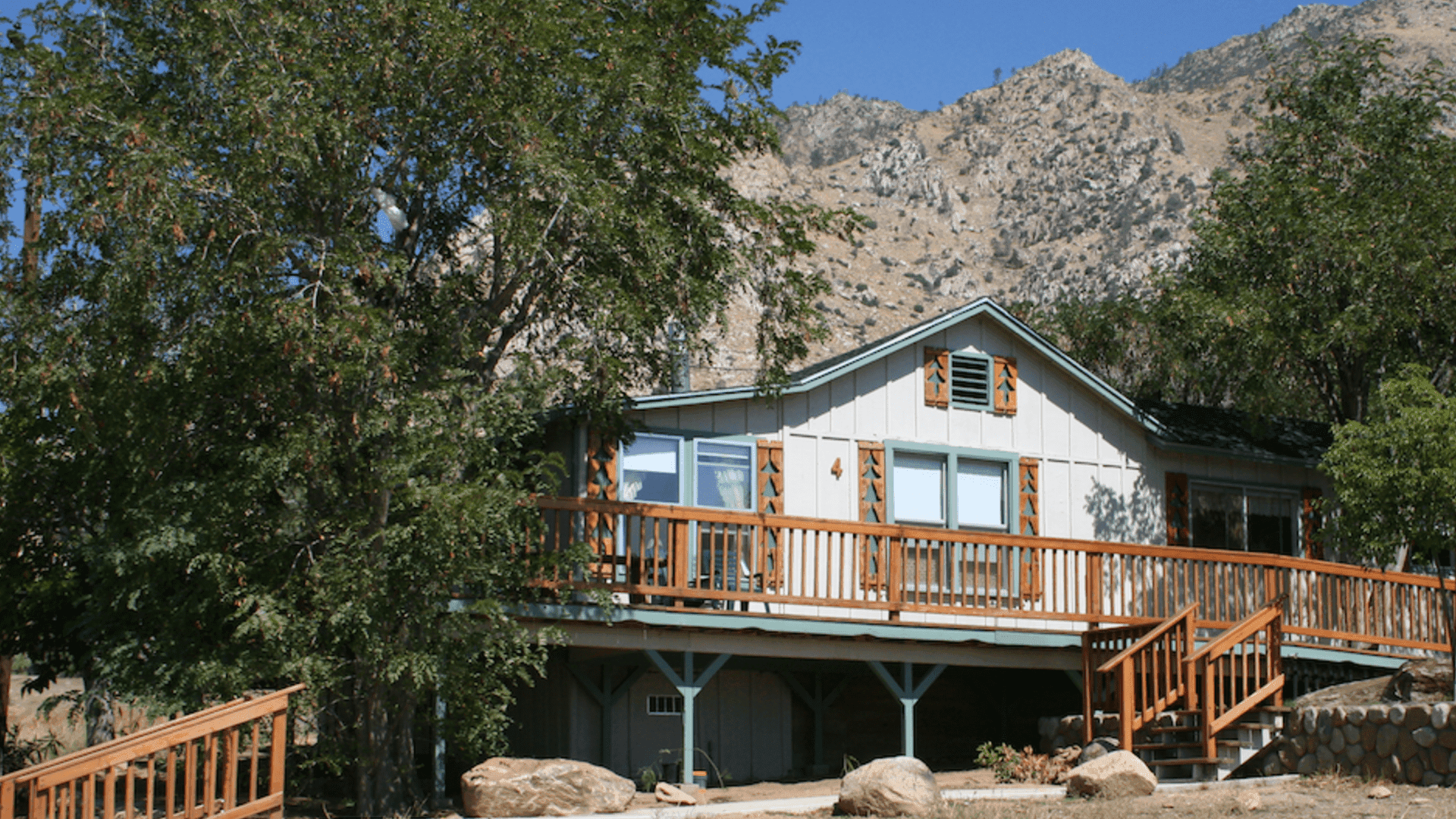 A rustic house with a wooden deck, surrounded by trees and mountains.