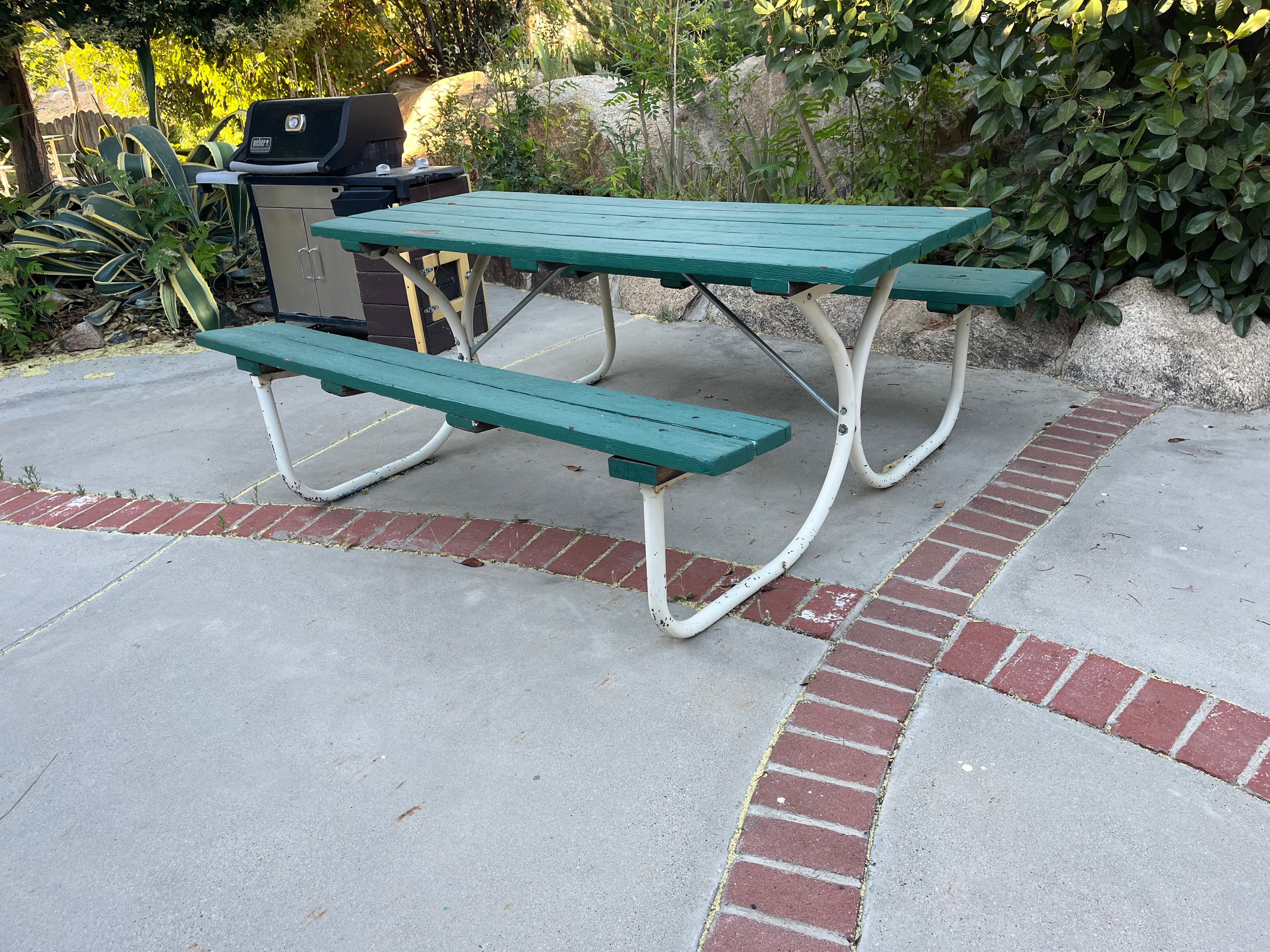 A green picnic table on a patio next to a grill and surrounded by plants.