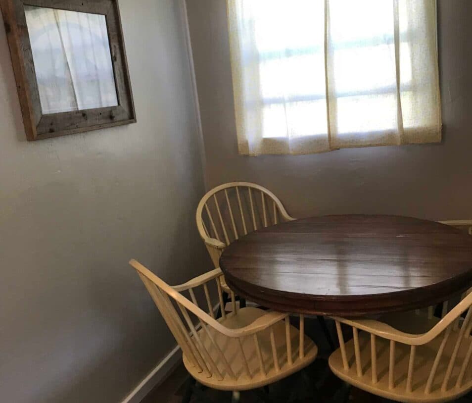 A small dining area with a round wooden table and two white chairs next to a window with light curtains.