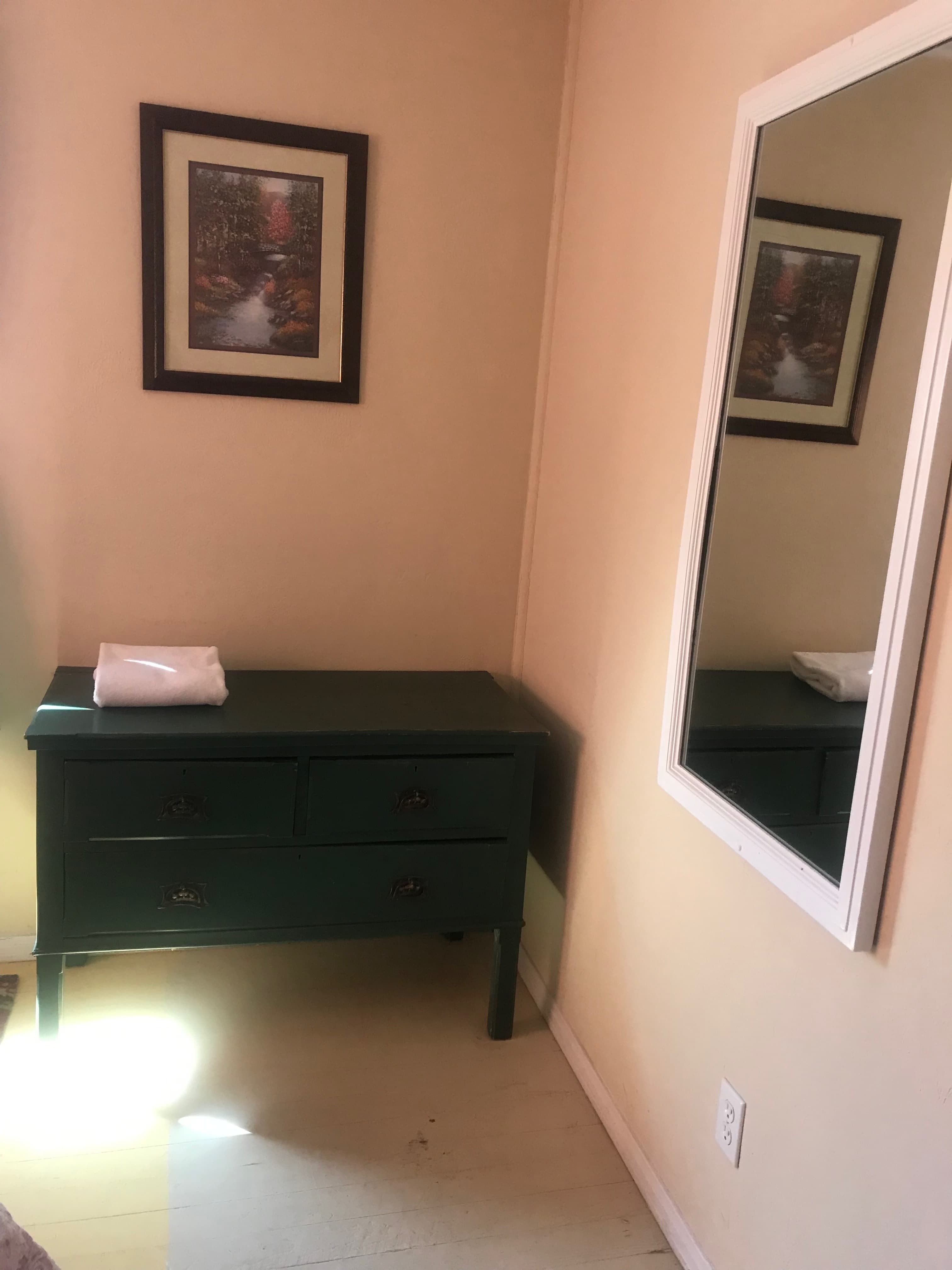 A green dresser with a white towel on top, next to a mirror and a framed landscape painting on the wall.