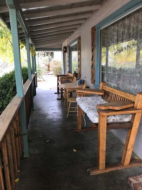 A covered porch featuring wooden furniture, including a bench and chairs, surrounded by greenery.