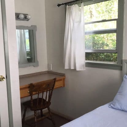 A small, bright room featuring a desk with a wooden chair, a mirror, and a bed with light blue bedding.