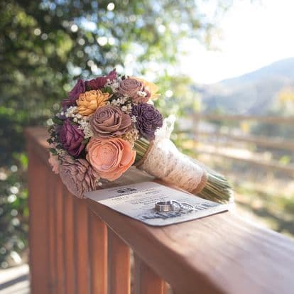 A colorful bouquet of flowers rests on a wooden railing beside a wedding ring and an invitation.