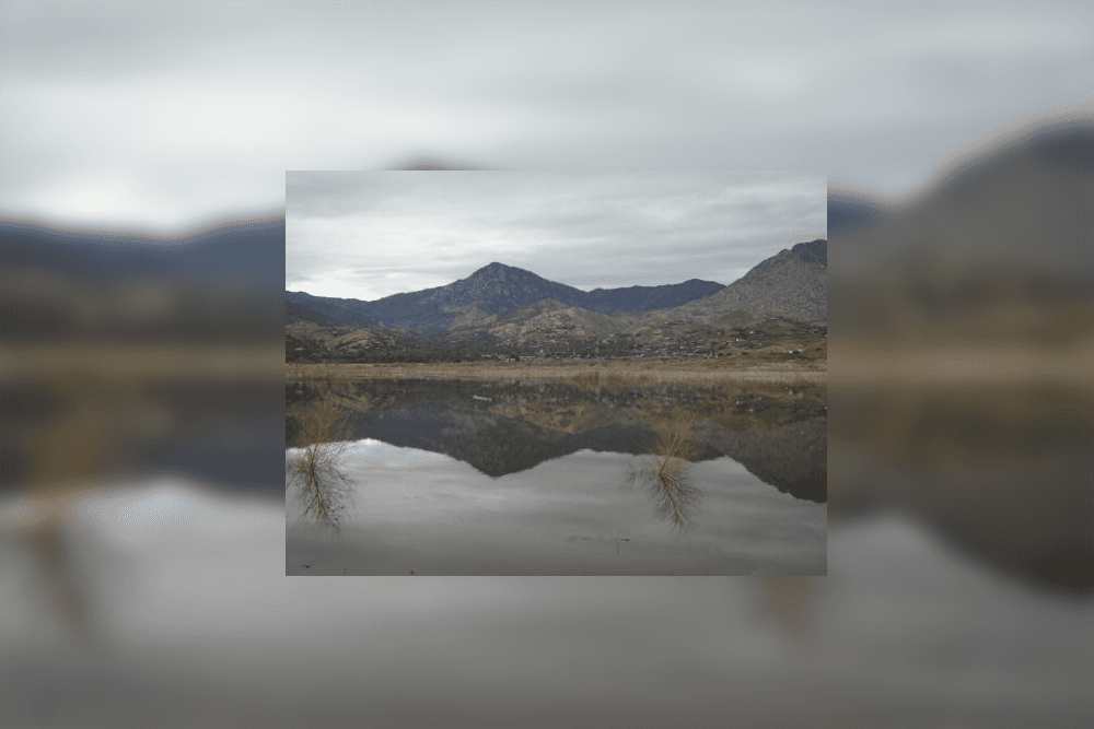 A calm lake reflects surrounding mountains under a cloudy sky.