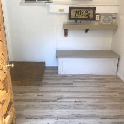 A small, partially furnished room featuring a welcome sign, a shelf, and a bench against a light-colored wall.