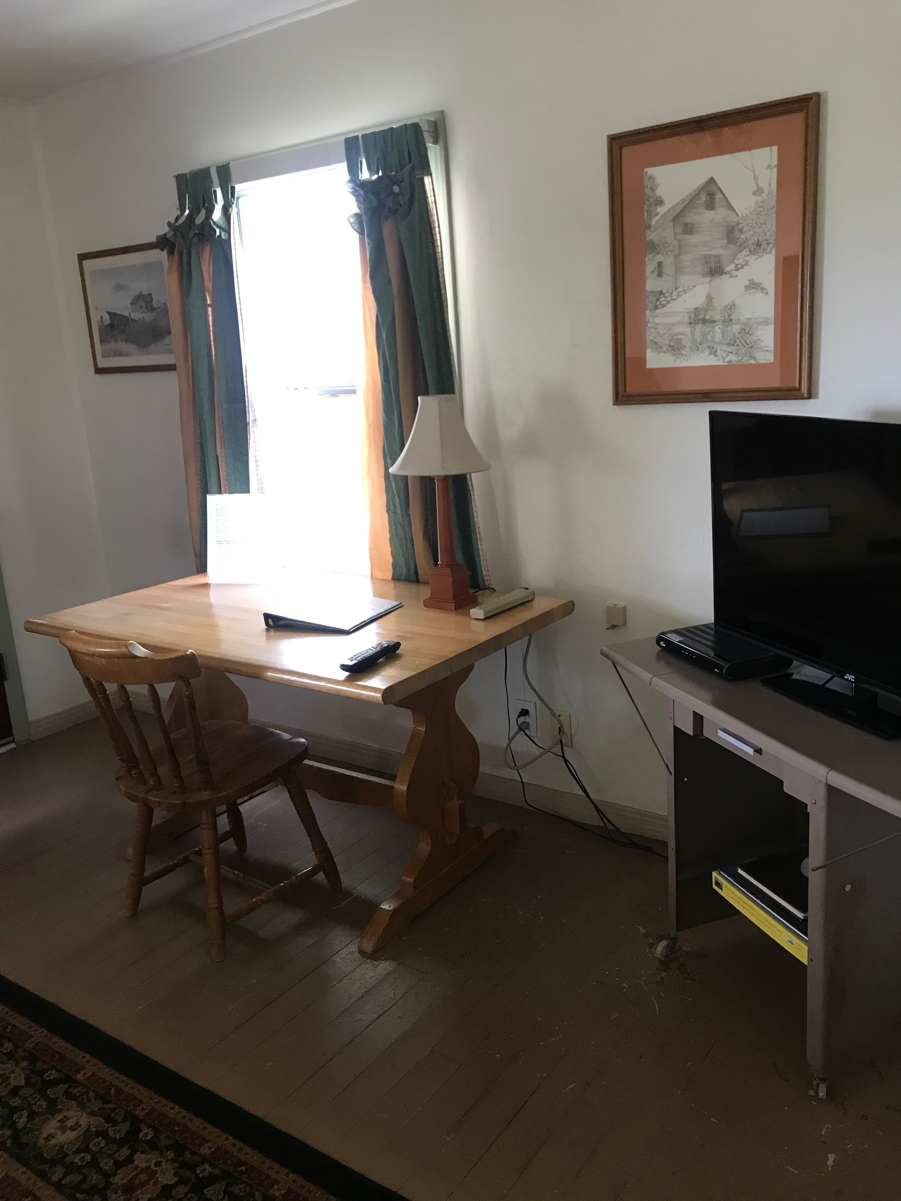 A wooden desk with a chair, a lamp, and a television stands in a room with curtains and a framed picture on the wall.